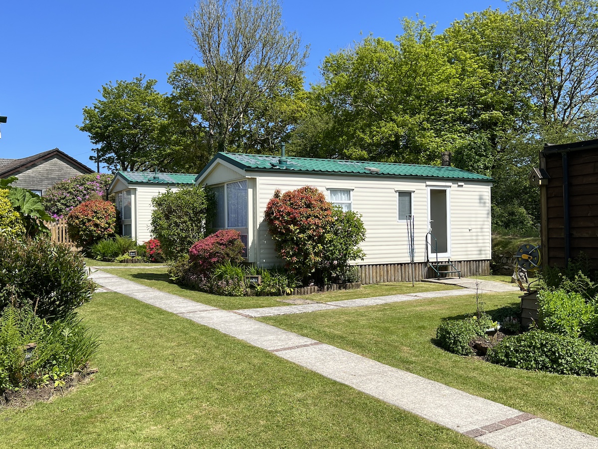 Cream caravan surrounded by multi-coloured plants and bushes, grass and concrete paths, partial view of another caravan on the left and corner of a wooden chalet on the right