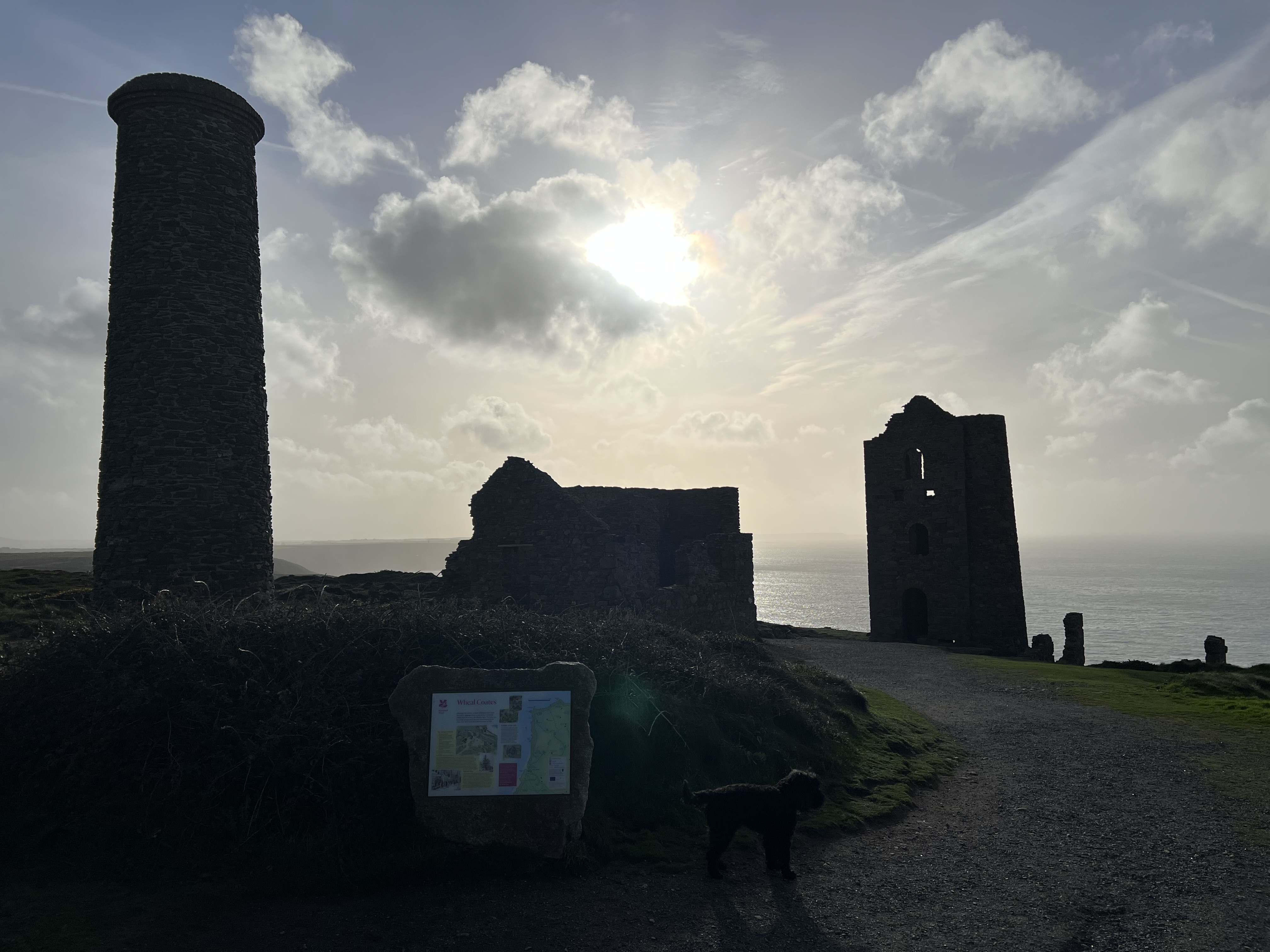 Old tin mine buildings in shadow with blue sky, clouds and sun in the background