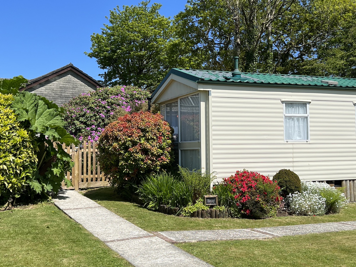 Cream caravan surrounded by multi-coloured plants and bushes, grass and concrete pathway with tall trees in the background