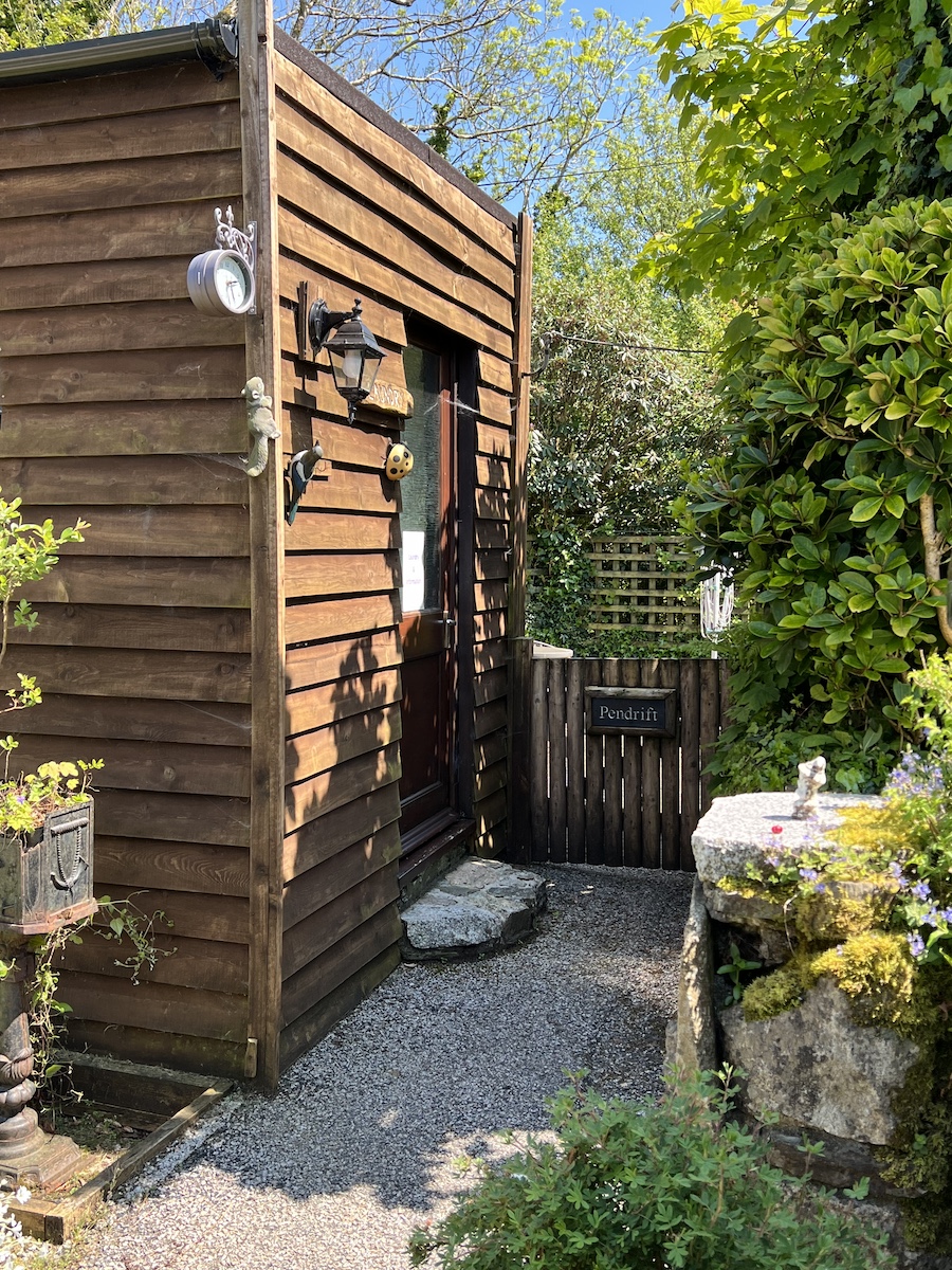 End of wooden chalet with front door, concrete pathway, wooden gate and plants/bushes