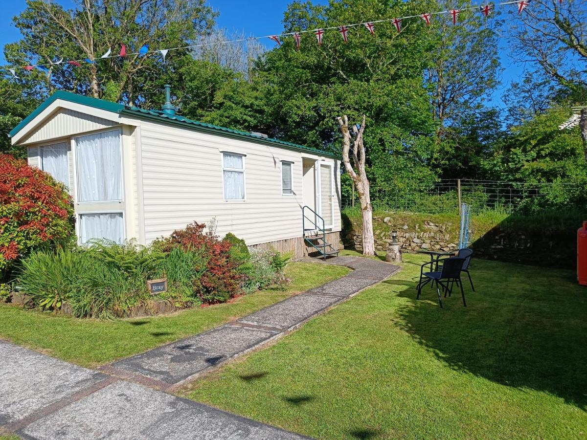 Cream caravan surrounded by green and red bushes, grass, concrete pathway and tall trees