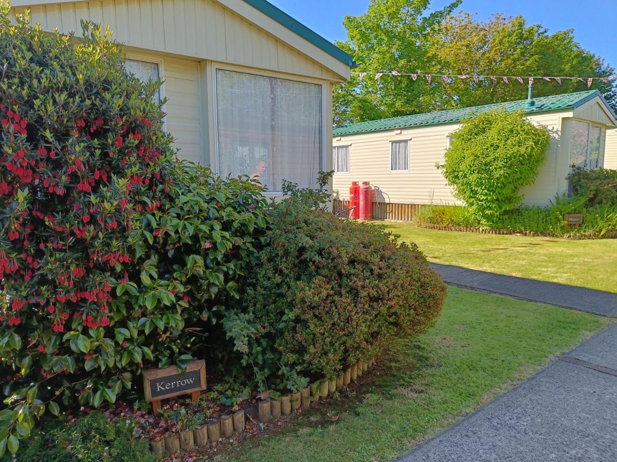 Green and red bushes with sign for 'Kerrow' in front of a cream caravan with another cream caravan, grass and bushes in the background