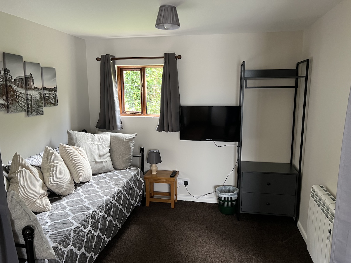 Room with white walls, brown flooring, sofa/day bed with grey and white cover and cushions, window, television on the wall and grey cupboard