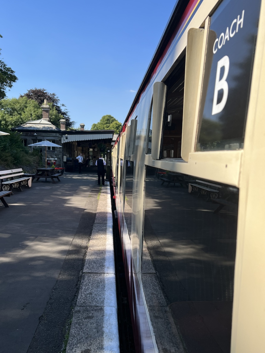 Black and cream train carriage with sign for 'Coach B' and concrete platform with station building in the background