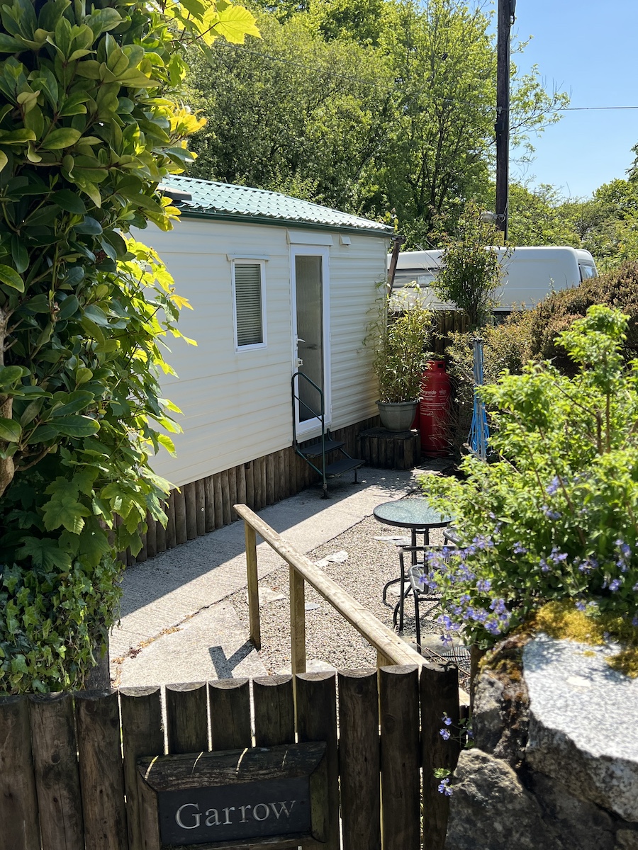 Wooden gate with sign saying 'Garrow' and cream caravan and plants